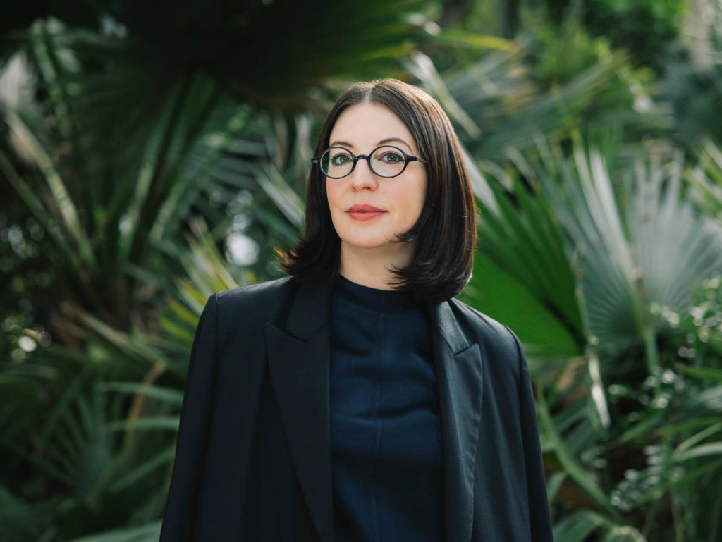 A person wearing a black blazer and dark shirt, standing in front of lush green palm leaves in a garden setting.