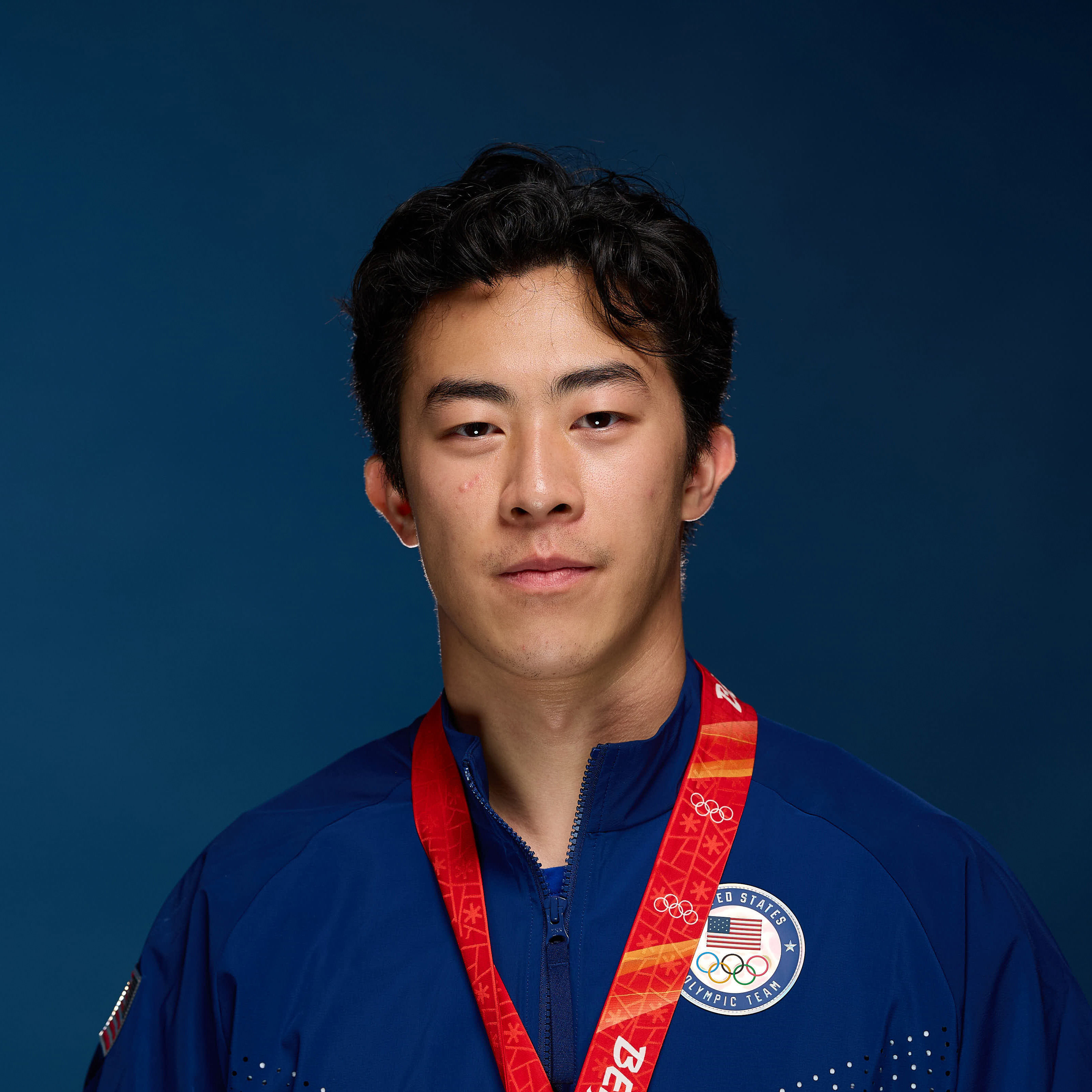 A person wearing a blue jacket with the United States Olympic Team logo, featuring a red medal ribbon against a solid blue background.
