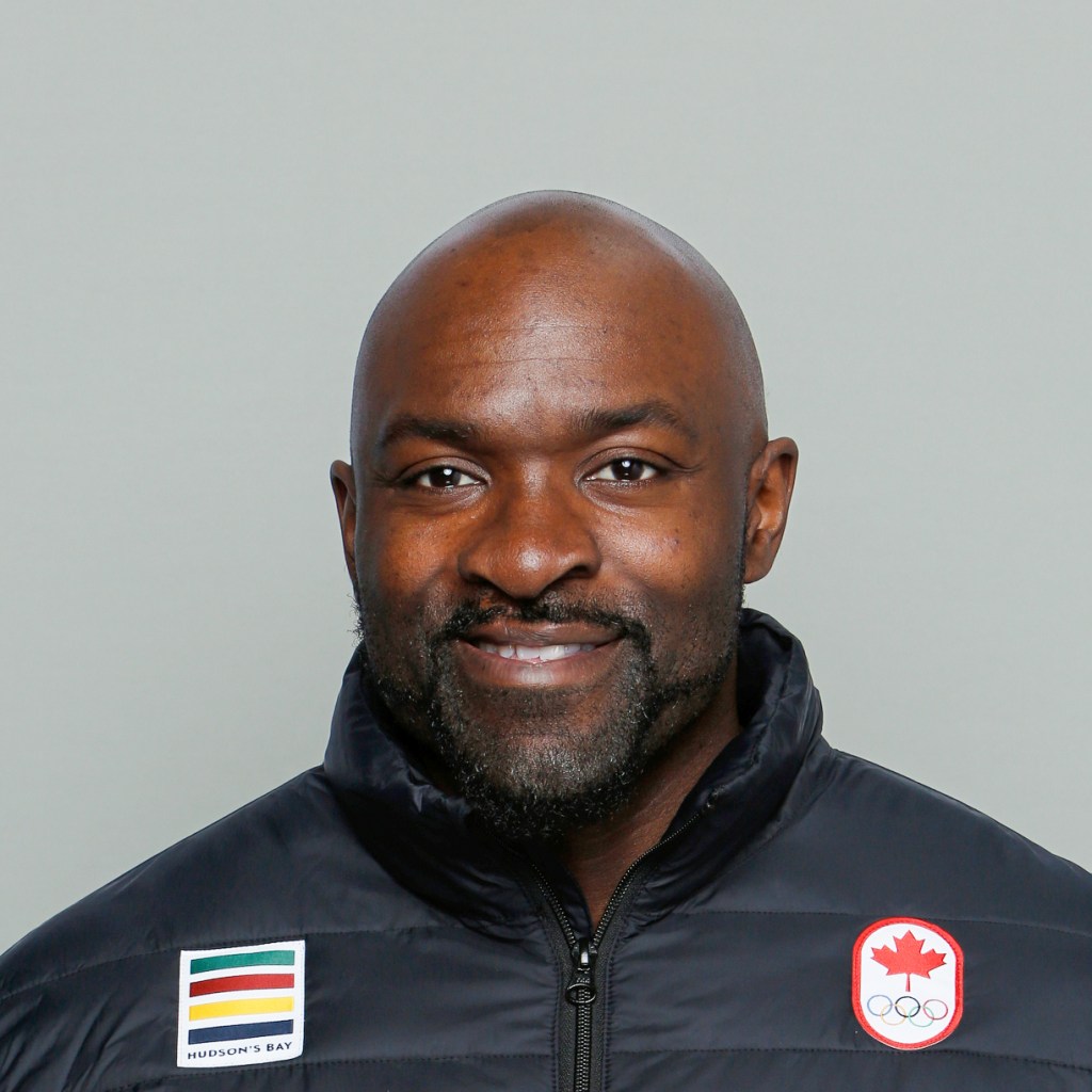 Close-up of an individual in a black jacket displaying a Canadian Olympic Team emblem with a red maple leaf, set against a neutral gray backdrop.