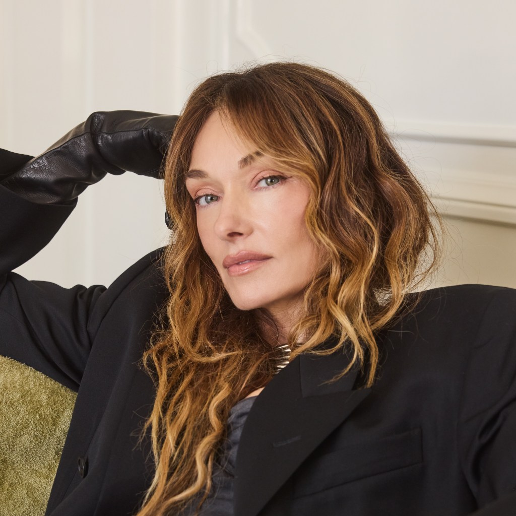 Person with long wavy brown hair wearing a black blazer and leather gloves, reclining on a green upholstered couch in a well-lit room.