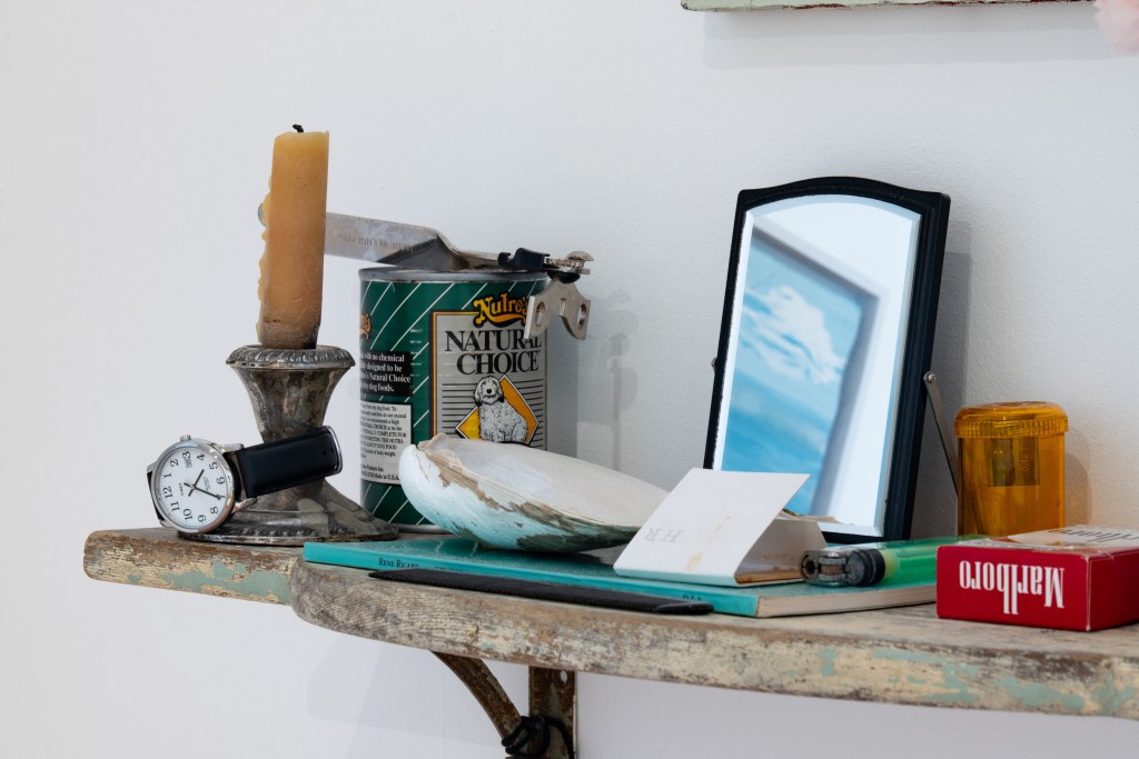 A vintage-inspired shelf arrangement showcasing a variety of items: a mirror reflecting blue sky, a candle, a tin labeled 'Natural Choice,' a watch, an oyster shell, a handwritten note, and more.