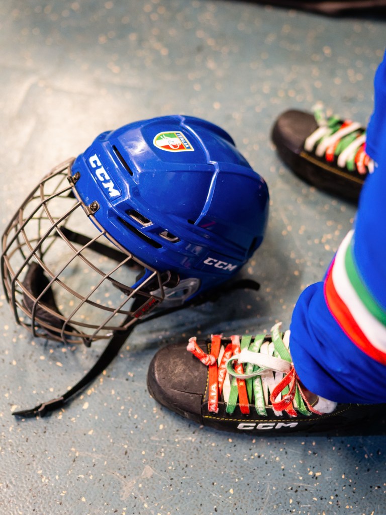 Close-up of a blue hockey helmet with an Italian flag logo next to a skate with laces in the colors of the Italian flag, on a speckled blue floor.