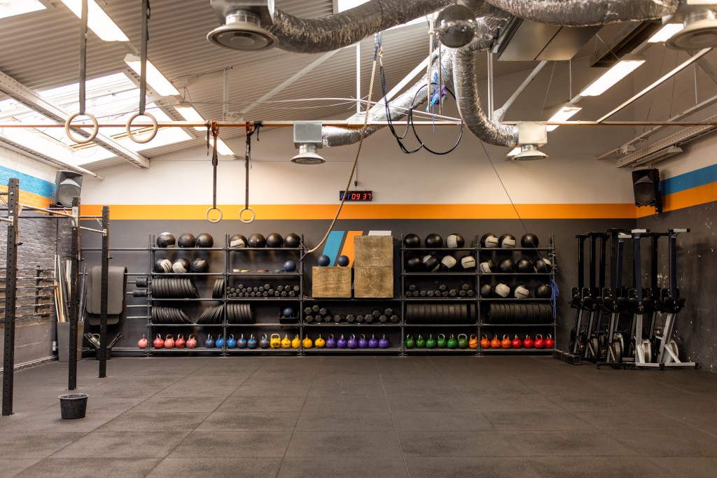 Interior of a brightly lit gym featuring a variety of fitness equipment such as kettlebells, medicine balls, weighted tires, pull-up rings, and rowing machines, with colorful accents on the walls.
