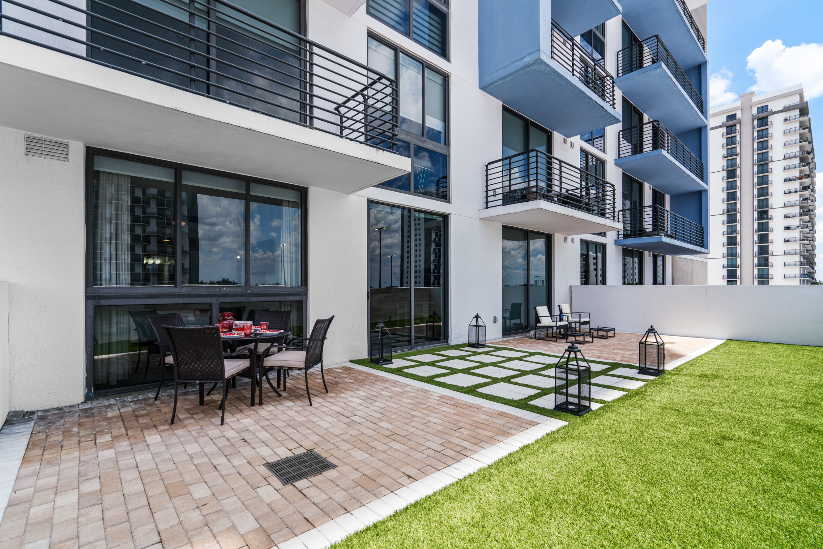 Outdoor patio area with a dining table set for four on a brick floor, green lawn with decorative lanterns, and a modern apartment building featuring balconies and large windows in the background.