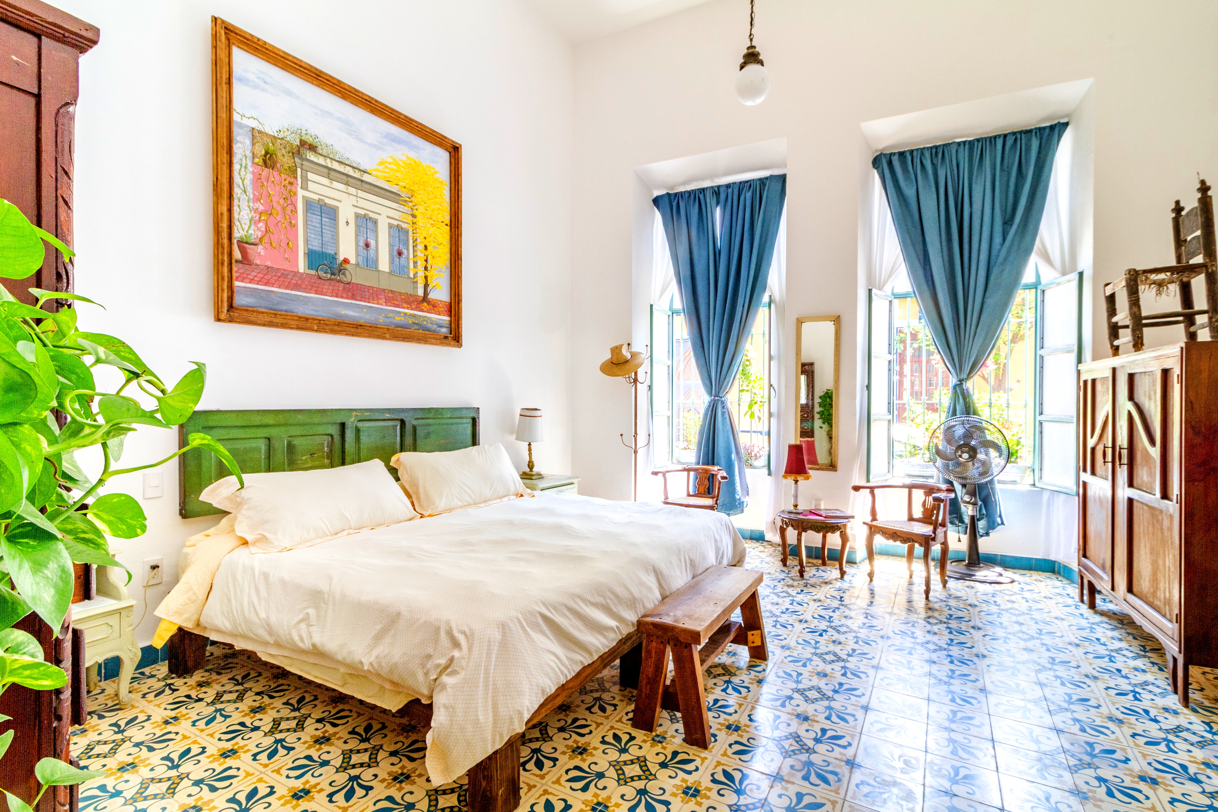 A vibrant bedroom showcases patterned tile floors, bright blue curtains, eclectic vintage furniture, and a colorful framed painting above a green-accented bed.