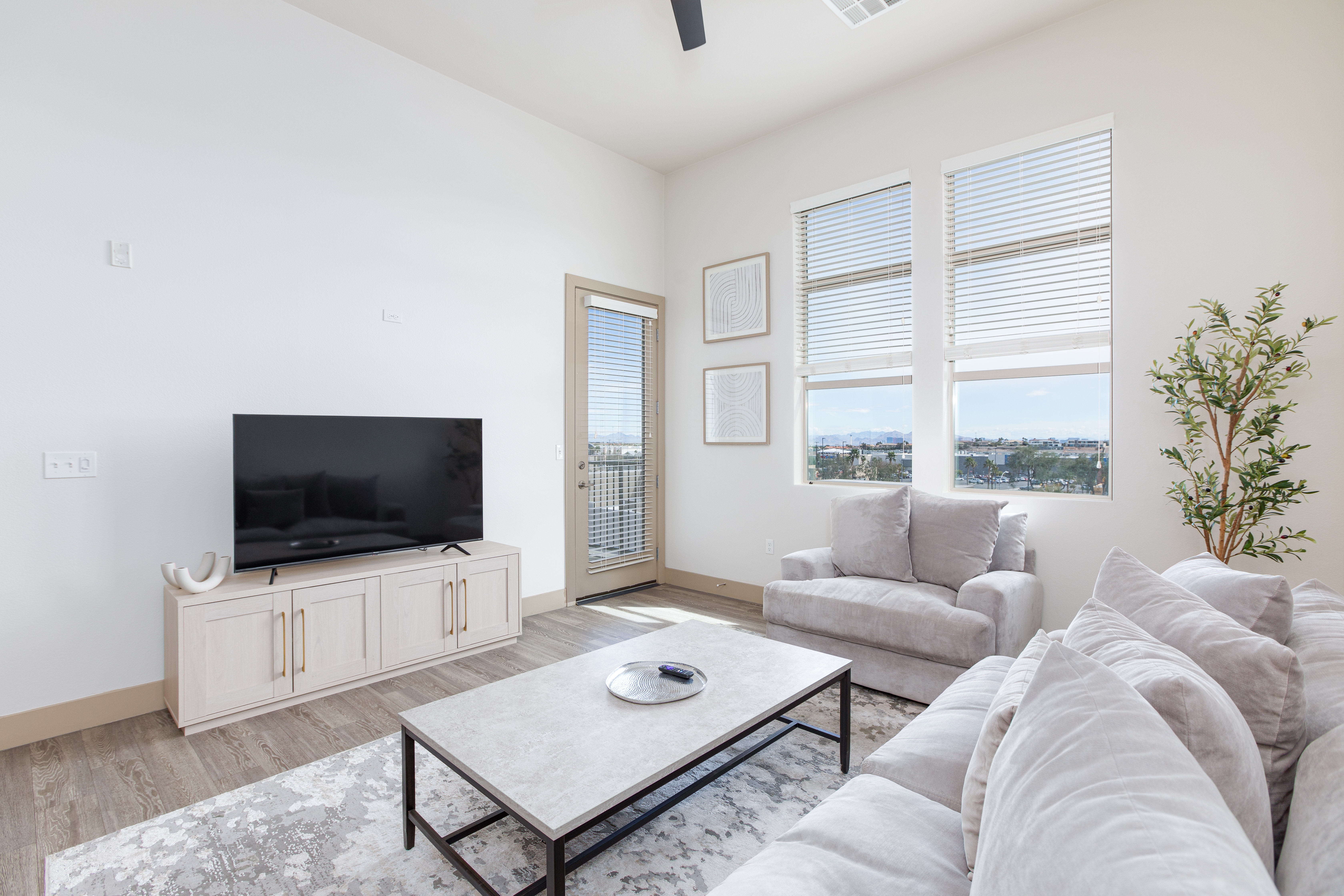 A bright modern living room features plush gray sofas, a large TV on a light wood console, expansive windows with city views, and minimalist decor.