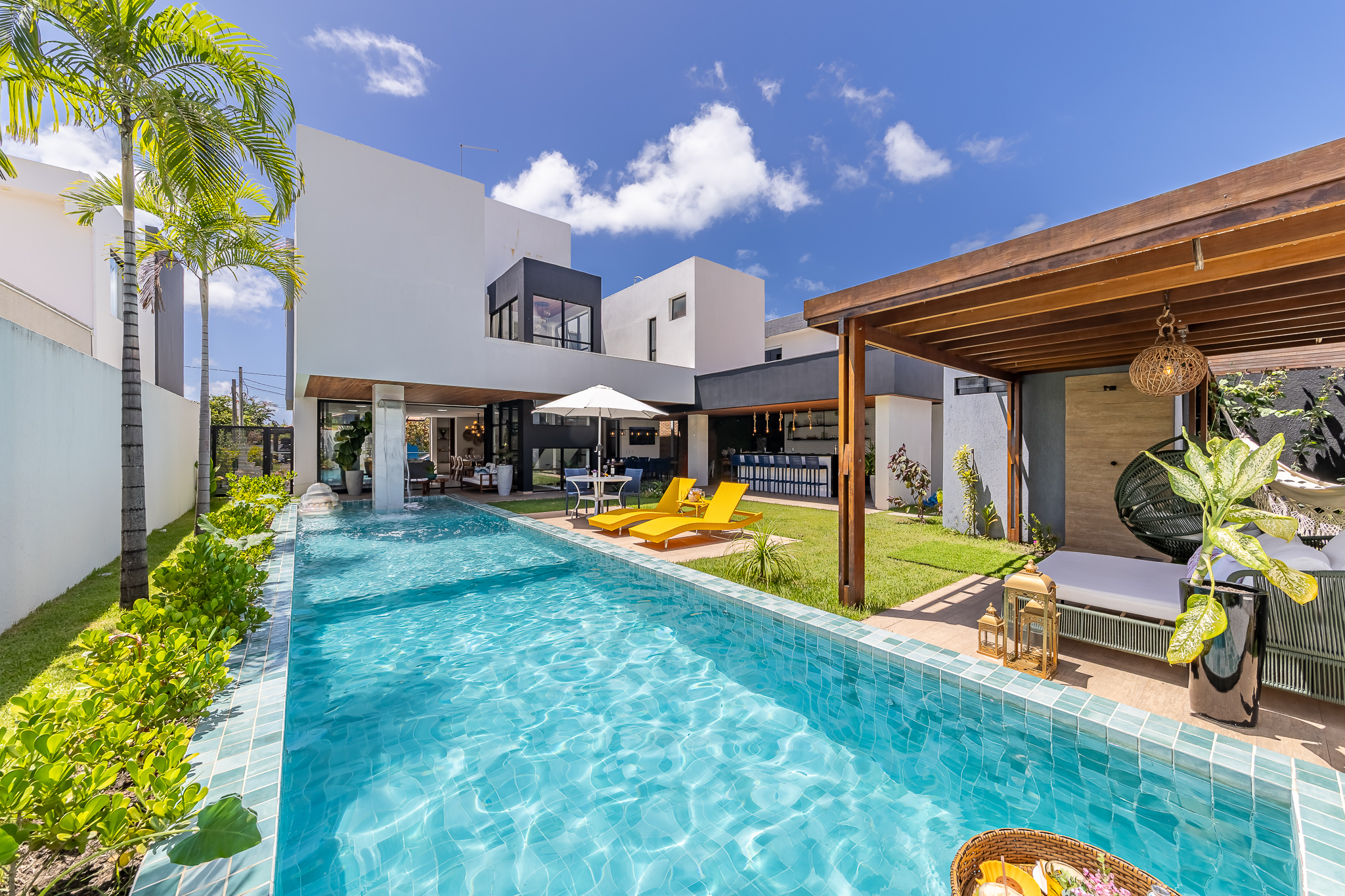 A contemporary villa boasts a sparkling turquoise pool surrounded by palm trees, sun loungers, and shaded outdoor lounges under a clear blue sky.