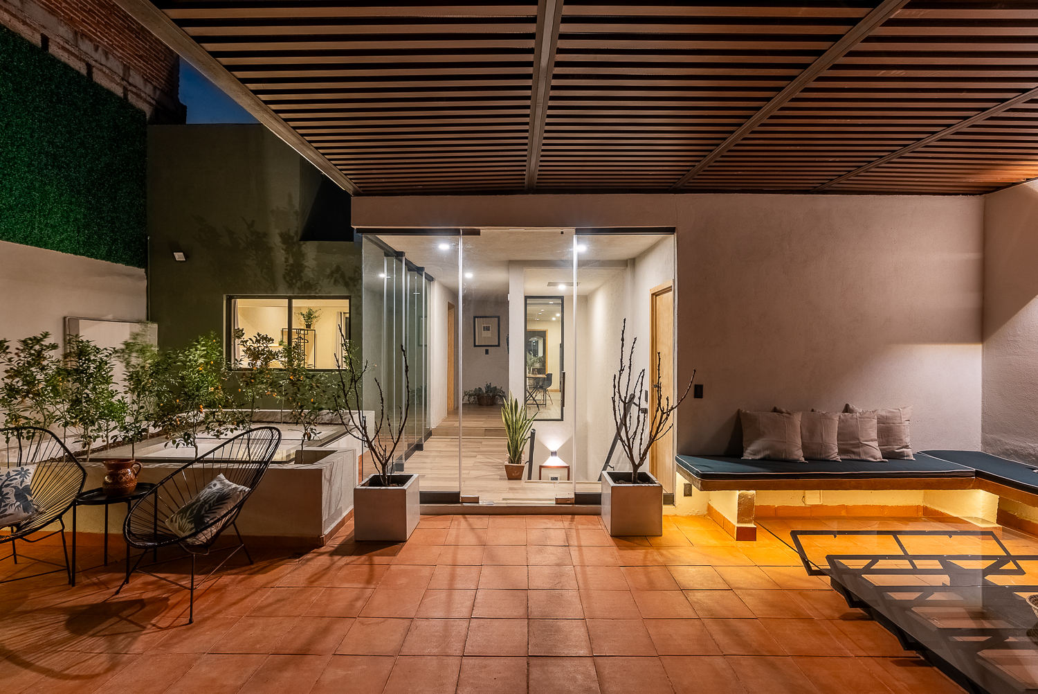 A welcoming patio space features modern outdoor seating, potted plants, and a built-in bench under a slatted roof, illuminated for evening comfort.