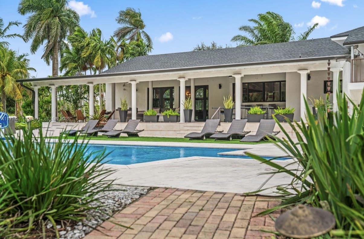 The outdoor area showcases a spacious patio with lounge chairs, a sparkling swimming pool, lush tropical landscaping, and a covered terrace supported by white columns.