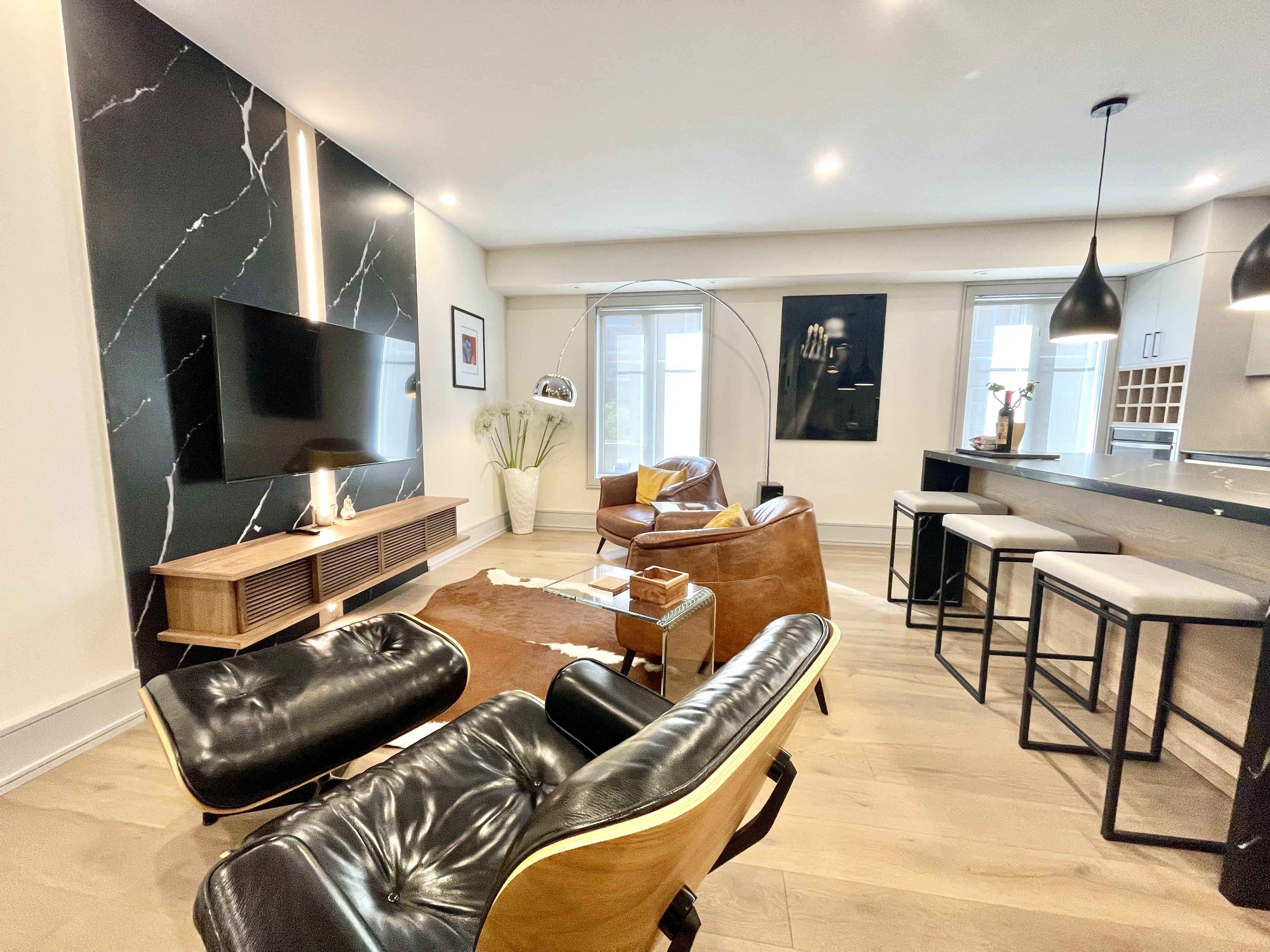The living room features modern d&eacute;cor with sleek black marble accents, tan leather chairs, a stylish lounge chair, and an open kitchen with bar stools under pendant lighting.