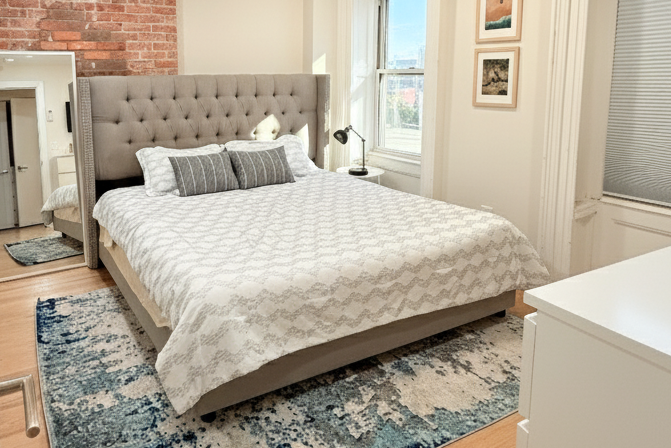 A bright, cozy bedroom features a tufted gray headboard, crisp white bedding, decorative pillows, and a blue patterned rug on hardwood floors.