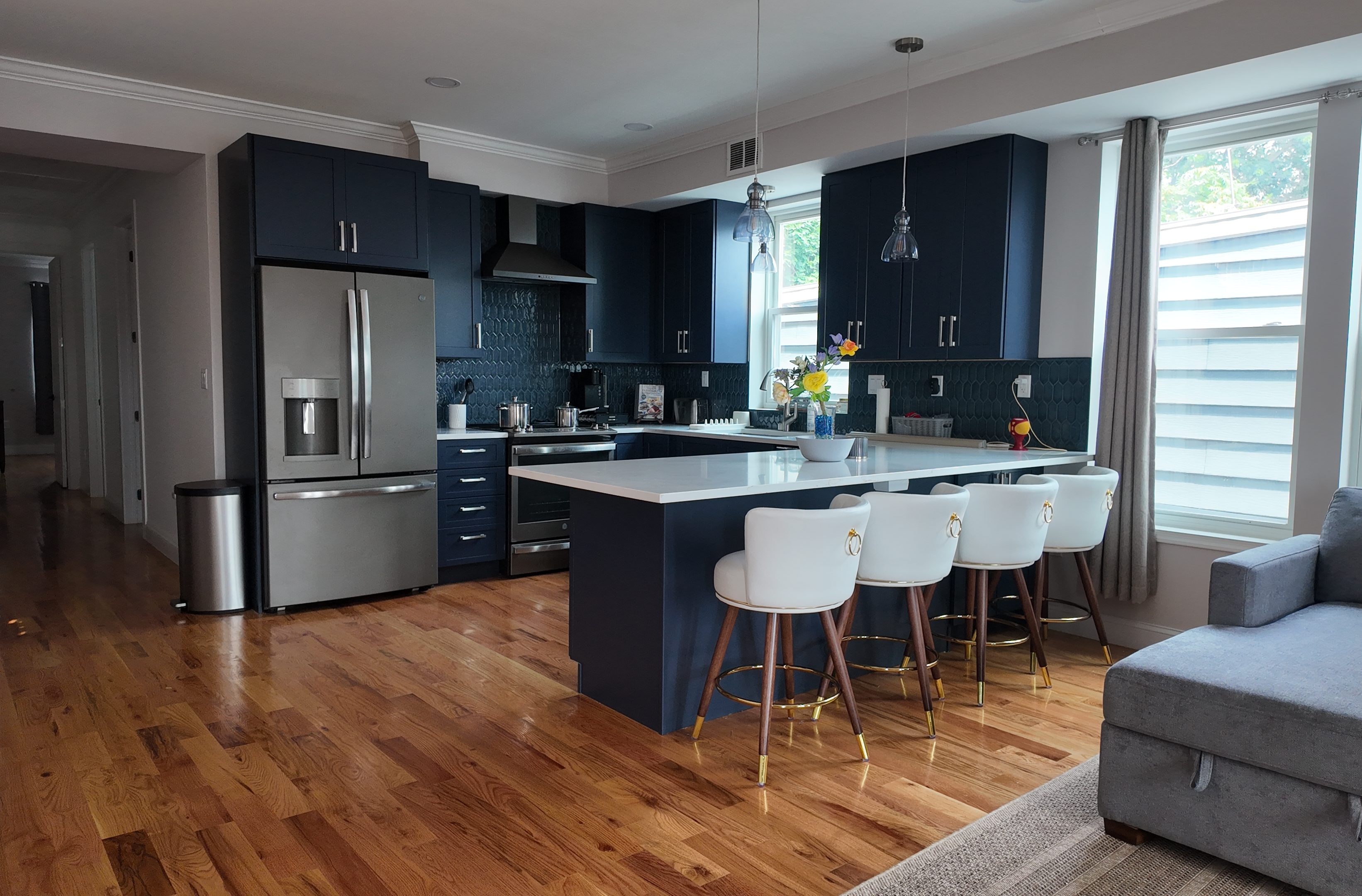 A modern kitchen with navy cabinets, stainless steel appliances, a large island with white countertops, and stylish white stools sits beside large windows letting in natural light.