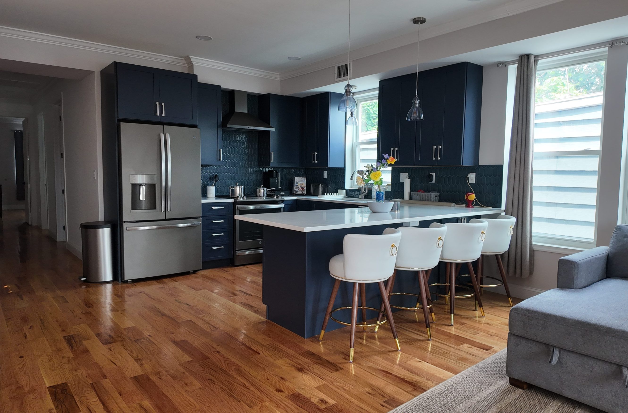 A modern kitchen with navy cabinets, stainless steel appliances, a large island with white countertops, and stylish white stools sits beside large windows letting in natural light.