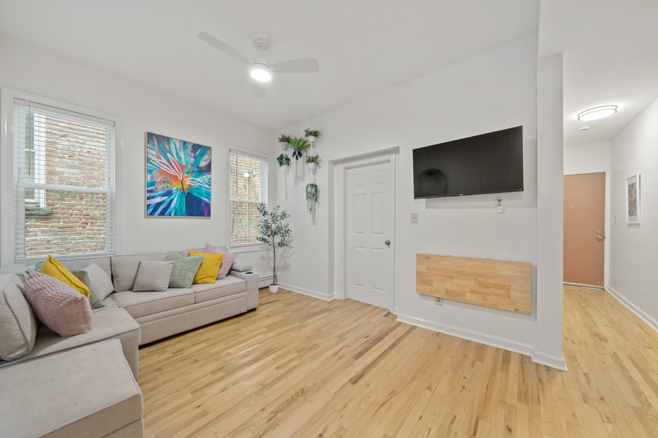 A cheerful living room with light wood floors, a beige sectional sofa adorned with colorful pillows, large windows, hanging plants, and vibrant wall art creates a welcoming vibe.