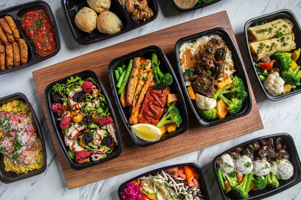 An assortment of healthy, gourmet meal prep dishes are neatly packed into black containers, each featuring a variety of proteins, vegetables, and grains.