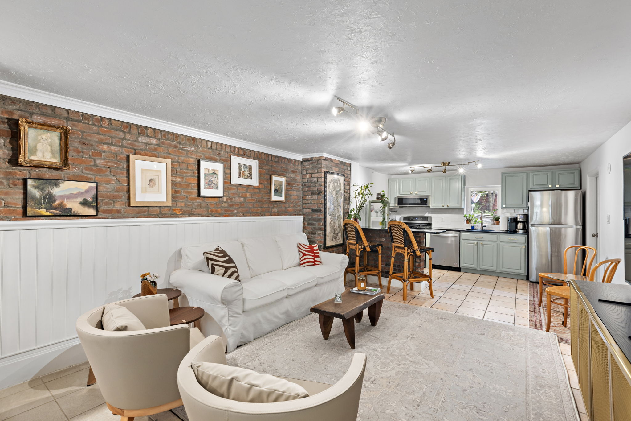 An inviting open-plan living space blends a white sofa, modern armchairs, and a kitchen with mint green cabinets, all set against a striking exposed brick wall decorated with artwork.