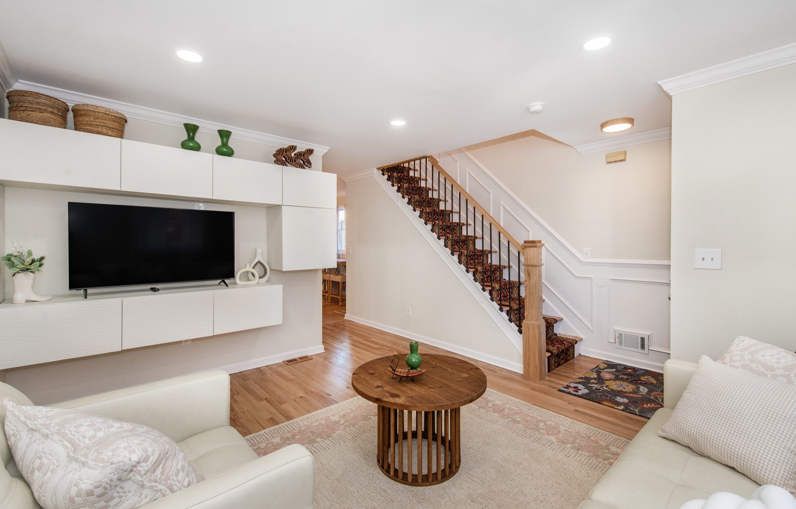 A stylish living room features cream-colored sofas, a sleek entertainment unit with a mounted TV, and a wooden staircase with decorative carpeting leading upward.