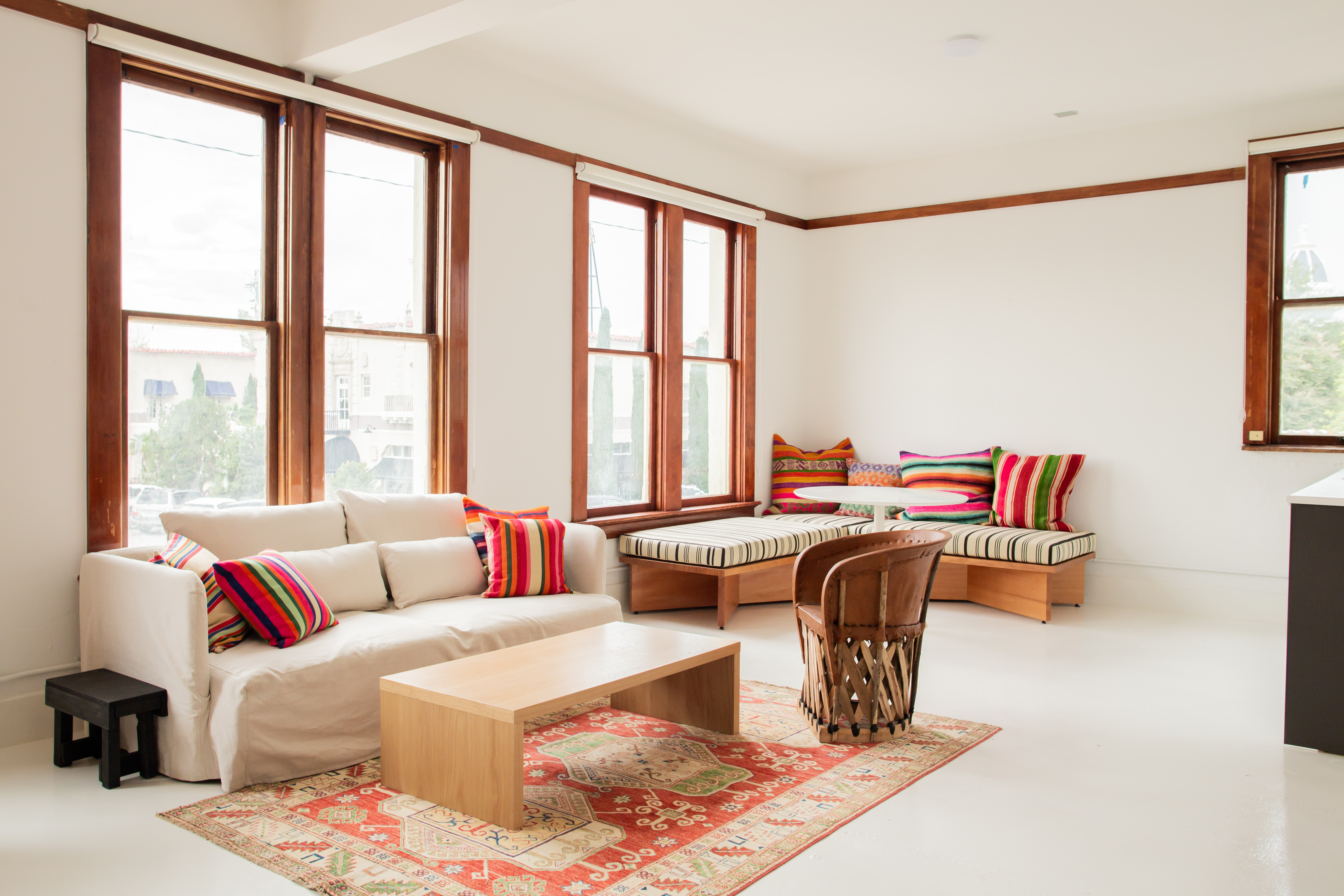 A bright, airy living space with white walls, rich wood-framed windows, colorful striped pillows, and eclectic Mexican-inspired furnishings.