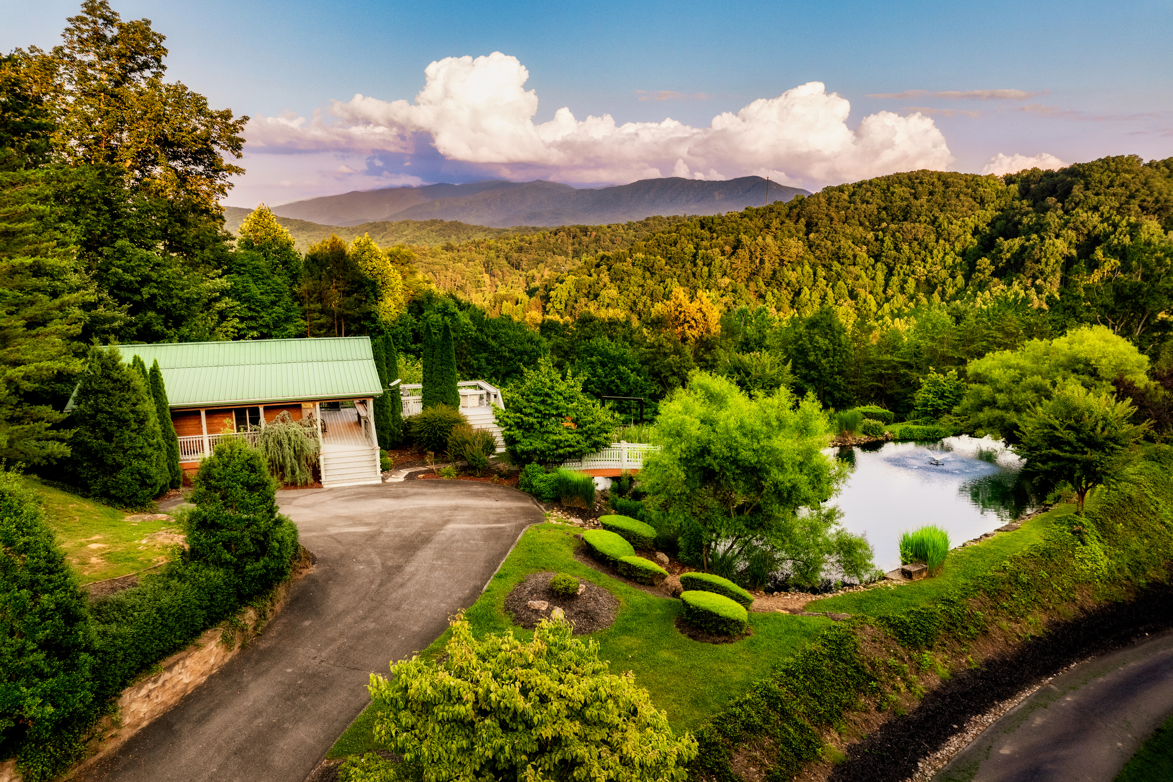 A charming green-roofed cabin nestled among manicured gardens and a serene pond sits against a sweeping panorama of lush Tennessee mountain forests under a dramatic summer sky.